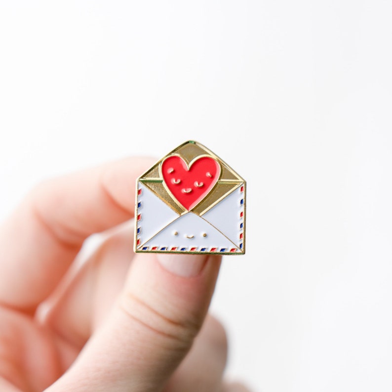 May include: Gold enamel pin shaped like an envelope with a red heart inside. The heart has a smiling face and the envelope has a smiley face.