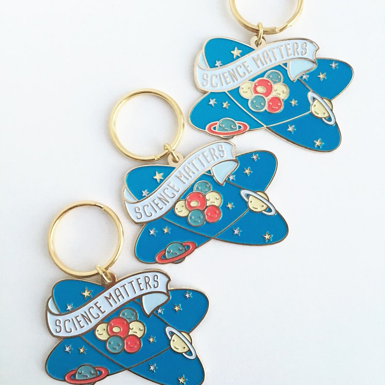 May include: Three gold keychains with blue and white enamel charms shaped like atoms. Each charm has a white banner with the text "Science Matters" and a cluster of colorful planets.