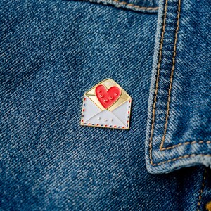 May include: A gold-coloured enamel pin shaped like an envelope with a red heart inside. The heart has a smiling face. The envelope has a white background with red and blue details.
