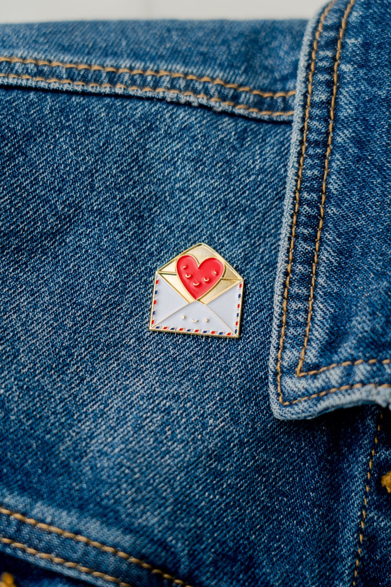 May include: A gold-coloured enamel pin shaped like an envelope with a red heart inside. The heart has a smiling face. The envelope has a white background with red and blue details.