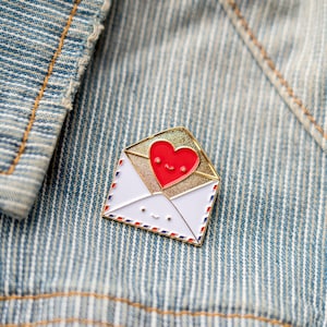 May include: A gold-coloured enamel pin shaped like an envelope with a red heart peeking out. The heart and envelope have smiling faces.