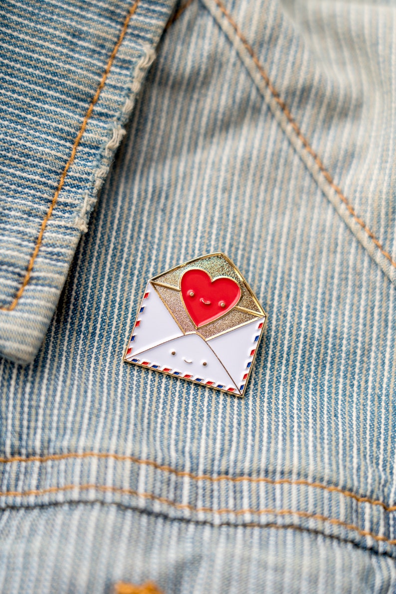 May include: A gold-coloured enamel pin shaped like an envelope with a red heart peeking out. The heart and envelope have smiling faces.