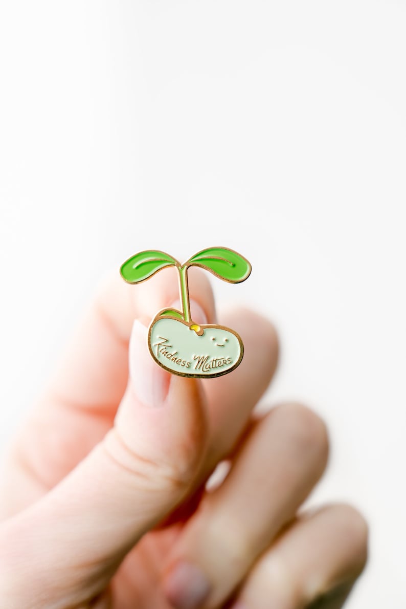 Pu&ograve; includere: Spilla in smalto dorato con un germoglio verde che cresce da un fagiolo verde chiaro. Il fagiolo ha un viso sorridente e il testo "Kindness Matters" scritto su di esso.