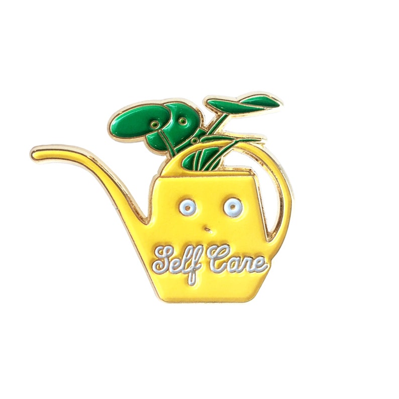 Puede incluir: Pin de esmalte amarillo de una regadera con una planta verde en su interior. La regadera tiene una cara con ojos y una sonrisa. El texto "Self Care" est&aacute; escrito en la parte delantera de la regadera.