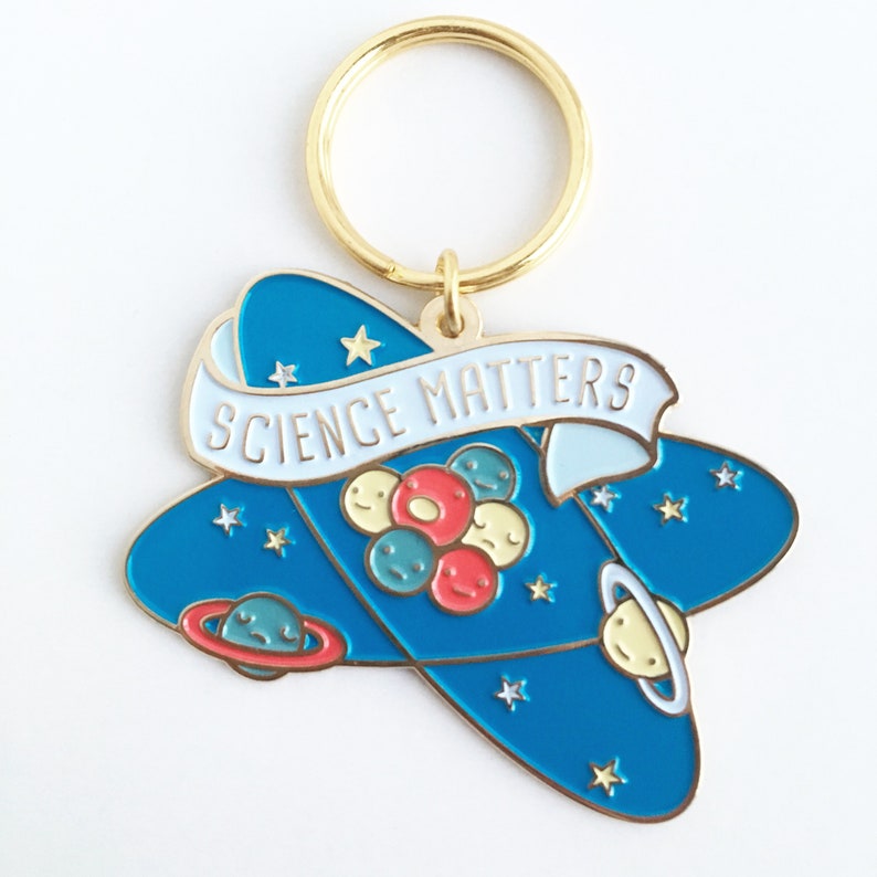 May include: A gold-colored keychain with a blue and white enamel design. The design features a stylized atom with planets orbiting around it. The text "SCIENCE MATTERS" is written on a white banner.