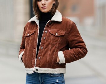 Può includere: Giacca di velluto a coste marrone con colletto e fodera in sherpa bianco. La giacca ha due tasche sul petto e due tasche laterali. La modella indossa jeans blu. La giacca ha un'etichetta rossa sulla tasca sul petto.