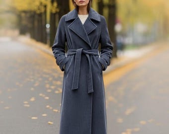 Puede incluir: Un abrigo largo de pana azul pizarra con cinturón envolvente. El abrigo presenta un cuello con muescas, mangas largas y bolsillos. La modelo está de pie en una carretera pavimentada con árboles al fondo.