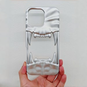 May include: Silver phone case with a stylized open-mouth design featuring sharp teeth. The case has a smooth, metallic finish and a cutout for the camera. The design resembles a ferocious animal's jaw.