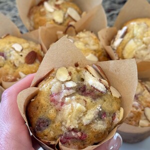 May include: A close-up shot of a muffin with almonds and red berries, presented in a brown paper case. The muffin is golden brown with visible berries and almond slices. Other muffins are in the background.