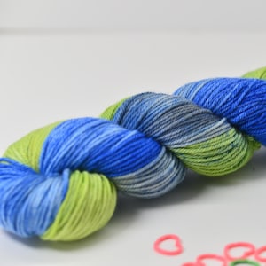 May include: A skein of hand-dyed yarn in shades of blue, green, and gray. The yarn is twisted into a compact bundle, with a few small pink and green heart-shaped stitch markers in the foreground. The yarn appears soft and textured.