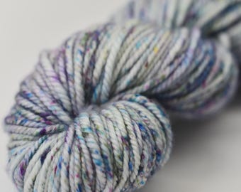 May include: Close-up of a skein of speckled yarn. The yarn is a mix of white, gray, purple, and blue fibers. The yarn is tightly wound and has a soft, textured appearance. Ideal for knitting or crochet projects.