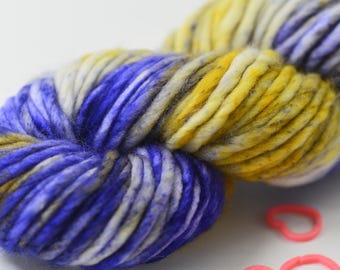 May include: Close-up of a skein of colorful yarn with shades of blue, yellow, and white. The yarn is thick and textured, with a speckled appearance. Pink and green heart-shaped rubber bands are scattered in the foreground.