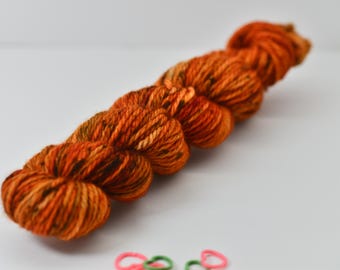 May include: A skein of orange and brown yarn, with a speckled appearance, lies on a white surface. The yarn is tightly wound, showcasing a blend of warm colors. Small pink and green rubber bands are at the bottom.