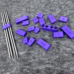 Puede incluir: Un conjunto de calibres y protectores de agujas de tejer morados. Los calibres están etiquetados con tamaños, y los protectores de agujas están unidos a un juego de agujas de tejer plateadas. Los artículos están colocados sobre una superficie gris.
