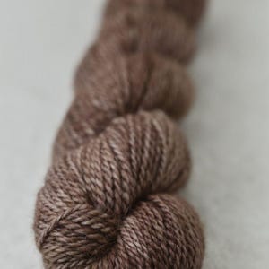 Puede incluir: Un ovillo de hilo marrón, bien enrollado y texturizado, con variaciones de color de marrón claro a oscuro. El hilo probablemente está hecho de fibras naturales, adecuado para proyectos de tejido o ganchillo.