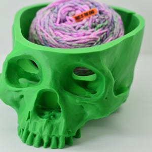 May include: A bright green skull-shaped container holding a ball of yarn. The yarn is a mix of pink, purple, and white. A small orange tag with the text "THIS IS THE END" is attached to the yarn. The container has eye sockets and a jaw.
