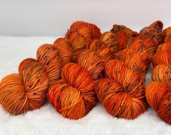 May include: A collection of multiple skeins of yarn in a vibrant orange hue, featuring subtle black speckles throughout the twisted fibers. The yarn is arranged in a cluster, showcasing the textured, multi-tonal appearance of the dyed material.