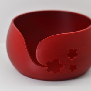 Può includere: Una ciotola per filato rossa con un'apertura curva e tre ritagli a forma di fiore. La ciotola è rotonda con una finitura liscia e opaca. Questa ciotola decorativa è progettata per contenere il filato durante la lavorazione a maglia o all'uncinetto.