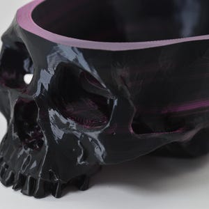 May include: A black skull-shaped bowl with a glossy finish and a pink interior rim. The skull has detailed features, including eye sockets, a nose cavity, and teeth. The bowl is likely made of a durable material.
