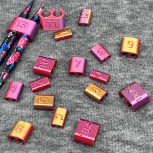 May include: A collection of knitting tools, including floral-patterned needles and stitch markers in various sizes. The stitch markers are in shades of pink, gold, and purple, with numbers indicating sizes.
