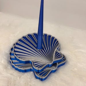 May include: A blue and silver ring holder featuring a base shaped like a scallop shell with a tall, slender, conical post extending upward from the center. The shell base has layered, ridged details in alternating blue and silver colors.