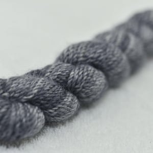 Puede incluir: Primer plano de un trozo de hilo gris, fuertemente retorcido en forma de cuerda. El hilo tiene un aspecto moteado con fibras grises claras y blancas intercaladas. El hilo se utiliza probablemente para proyectos de punto o ganchillo.