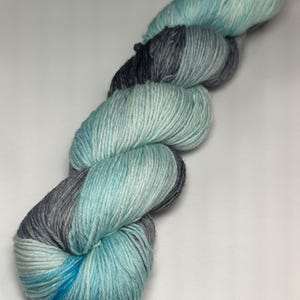 May include: A skein of hand-dyed yarn in shades of teal, gray, and blue. The yarn is twisted and shows a gradient effect, transitioning from darker gray to lighter teal and blue hues. The yarn is likely for knitting or crocheting projects.