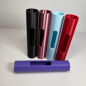 May include: A collection of five cylindrical objects in various colors: black, pink, light blue, red, and purple. Each has a rectangular cutout. The objects are arranged on a white surface, with four standing upright and one lying horizontally.