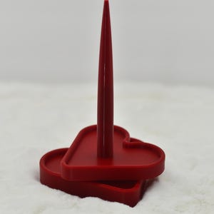 May include: A red heart-shaped candle holder with a tall, pointed center. The holder has a two-tiered design, with a base and a top section, both in the shape of a heart. The candle holder is made of a solid, smooth material.
