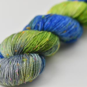 May include: Close-up of a skein of yarn in shades of blue, green, and gray. The yarn has a speckled appearance with flecks of orange and pink. The yarn is likely used for knitting or crocheting projects.