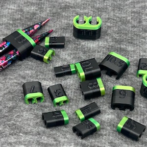 Puede incluir: Un conjunto de calibres y protectores de agujas de tejer negros y verdes. Los calibres están etiquetados con tamaños, y los protectores están diseñados para adaptarse a las agujas. Las agujas tienen un estampado floral.