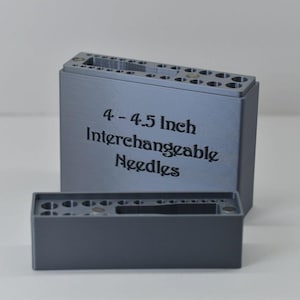 Peut inclure: Deux supports d'aiguilles rectangulaires gris. Le plus grand porte le texte "4 - 4.5 Inch Interchangeable Needles". Le plus petit est devant. Les deux ont plusieurs trous pour les aiguilles.