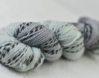 Puede incluir: Primer plano de varios ovillos de hilo en diferentes tonos de gris y azul, con motas negras. El hilo está dispuesto en una fila, con el foco en el primer ovillo. El hilo parece suave y esponjoso, ideal para proyectos de punto o ganchillo.