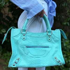May include: A turquoise leather handbag with a snakeskin pattern. The bag has a top handle, a front zipper pocket, and silver-tone hardware. The bag is held by a person wearing a light blue shirt.