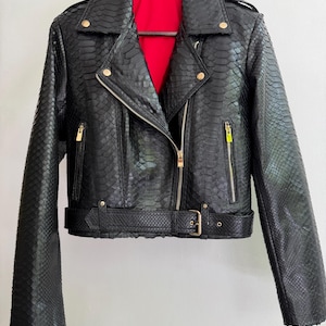 Puede incluir: Chaqueta de moto de cuero sintético negro con textura de piel de serpiente, cremalleras doradas y un interior rojo. La chaqueta presenta un cuello con solapas, un cinturón en la cintura y un estilo motero clásico.