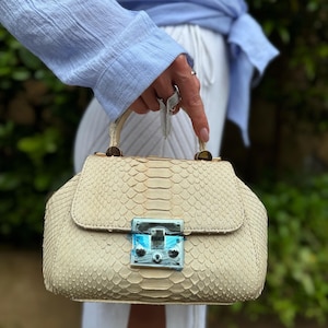 Puede incluir: Bolso de mano color crema con un patrón de piel de serpiente texturizado. El bolso tiene un cierre de solapa con un cierre plateado y un asa corta. El bolso lo sostiene una persona que lleva una camisa de manga larga azul claro.