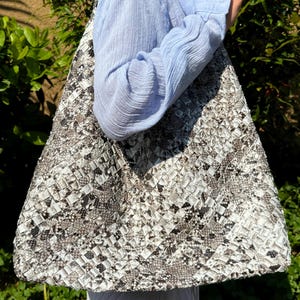 Può includere: Una grande borsa tote con un motivo intrecciato nei toni del grigio, bianco e nero. La borsa ha una parte superiore ampia e aperta e una tracolla. La borsa è realizzata in un materiale strutturato e sembra robusta. La borsa viene trasportata.