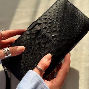以下が含まれることがあります： 黒色の財布。鱗のような模様のテクスチャが施されています。長方形の財布にはジッパーが付いています。画像には、2つの手で財布が持たれており、指にはシルバーの指輪がはめられています。
