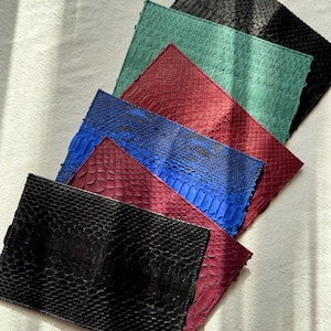 Op de afbeelding: Een verzameling handgemaakte clutches in verschillende kleuren, waaronder zwart, blauwgroen, bordeauxrood en blauw. De clutches zijn gemaakt van een textuurrijk, op slangenleer lijkend materiaal. De clutches zijn in een waaier gerangschikt, met de zwarte clutch op de voorgrond.
