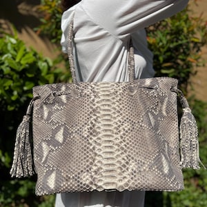 May include: A large, rectangular tote bag with a snakeskin pattern in shades of grey and cream. The bag has a drawstring closure and matching tassels on each side. The handles are also made of the snakeskin material.