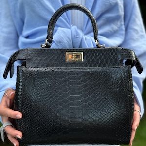 Op de afbeelding: Een zwarte handtas met een textuur van slangenleer. De tas heeft een handvat aan de bovenkant, een rechthoekige vorm en een goudkleurige sluiting. De tas wordt vastgehouden voor een persoon die een lichtblauw overhemd draagt.