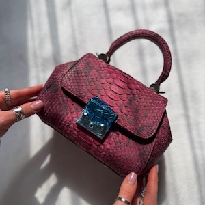 May include: A burgundy handbag with a snakeskin pattern. The bag has a top handle and a flap closure with a blue, jewel-like clasp. The bag is held by two hands, showcasing the bag's design and color.