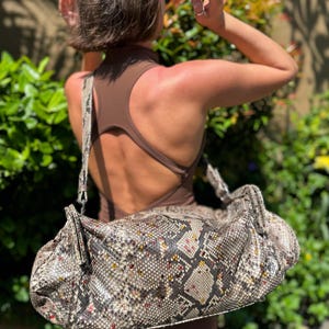 Puede incluir: Un bolso grande y texturizado con un estampado de piel de serpiente en tonos grises, marrones y crema. El bolso tiene una correa para el hombro y se lleva sobre el hombro. El bolso es el foco de la imagen.
