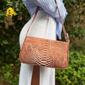 Puede incluir: Un bolso rectangular de color cobre con un estampado de piel de serpiente. El bolso tiene una correa fina a juego y una hebilla plateada. El fondo presenta follaje verde y flores amarillas.