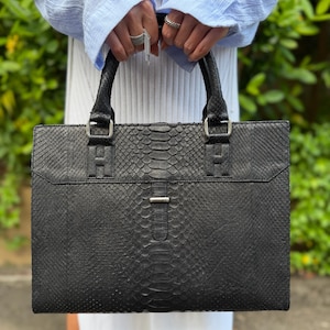May include: A black rectangular handbag with a snakeskin texture. The bag features two handles, silver-tone metal hardware, and a flap closure. The bag is held by a person wearing a light blue shirt and rings.