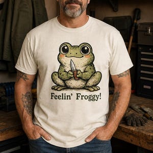 Może przedstawiać: Kremowy t-shirt z kr&oacute;tkim rękawem z grafiką zielonej żaby trzymającej mały n&oacute;ż. Pod żabą znajduje się napis "Feelin' Froggy!" w ciemnobrązowym kolorze. Koszulka posiada okrągły dekolt i standardowy kr&oacute;j.