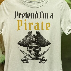 May include: White t-shirt with the text "Pretend I'm a Pirate" in black and gold. The shirt features a skull wearing a pirate hat with crossed swords. The design is centered on the front of the shirt.