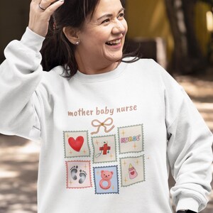 K&ouml;nnte beinhalten: Wei&szlig;es Sweatshirt mit dem Text "mother baby nurse" und mehreren Briefmarken-Grafiken. Die Grafiken zeigen ein rotes Herz, ein Kreuz, das Wort "nurse", Babyf&uuml;&szlig;e, einen Teddyb&auml;r und eine Babyflasche.