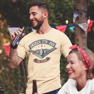 Puede incluir: Una camiseta de manga corta color crema con un estampado gr&aacute;fico de un avi&oacute;n bombardero furtivo sobre el texto AMERICA 1776-2026 y 250 YEARS, enmarcado por un dise&ntilde;o de pancarta con estrellas y una est&eacute;tica vintage desgastada.