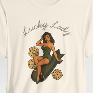 May include: A cream-colored short-sleeve t-shirt featuring a graphic illustration of a person in a green swimsuit sitting on a dark bomb, surrounded by three dice. The text Lucky Lady is printed in a cursive font above the central illustration.